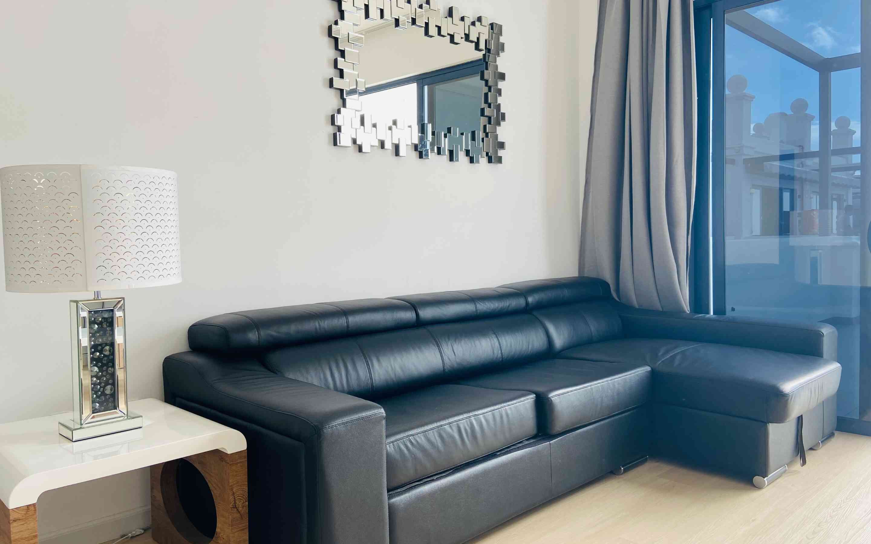 A modern living room with a black leather sofa, a white side table, a decorative mirror, and a large window with curtains.