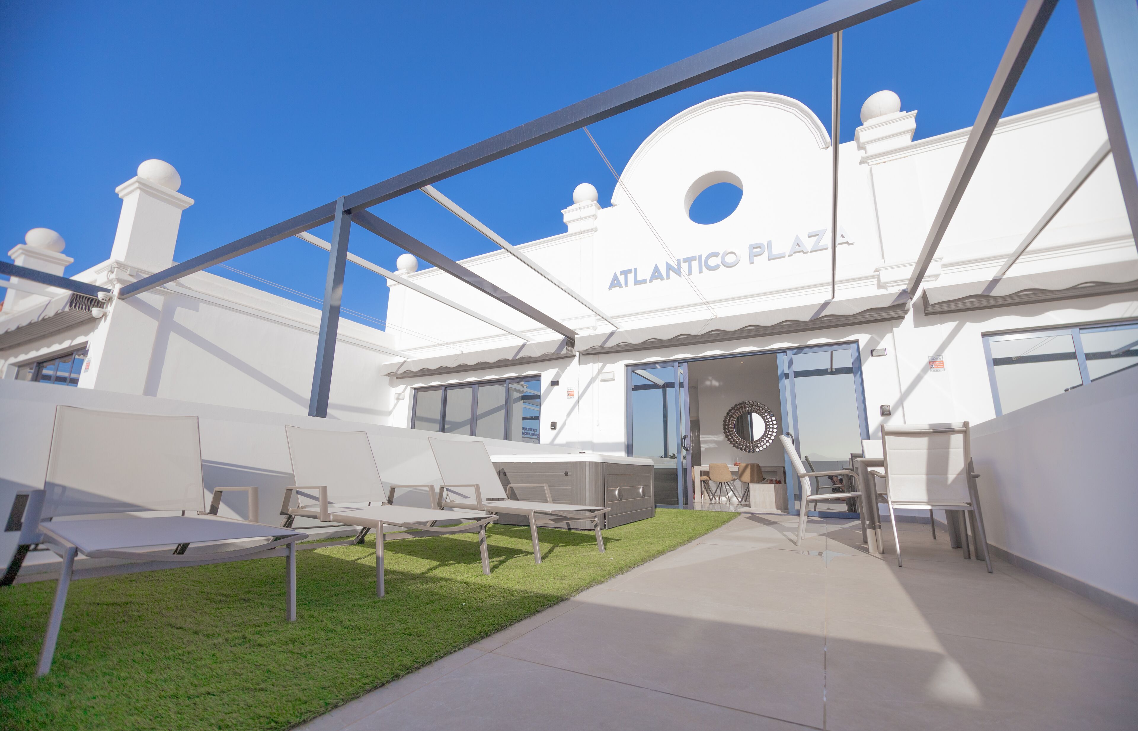 Un espace extérieur aménagé avec des chaises blanches et une petite table, situé devant un bâtiment portant l’inscription « ATLANTICO PLAZA ».