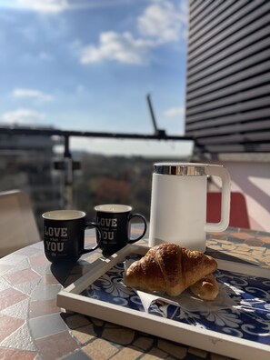 Une table sur laquelle se trouve un plateau contenant un croissant et deux tasses, l’une avec un motif et l’autre unie, posée sur une table carrelée avec vue sur le ciel et un bâtiment en arrière-plan.