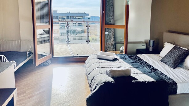A modern hotel room with a large bed, a small desk, and a balcony with a city view.