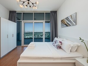 A modern bedroom with a large bed, a city view through floor-to-ceiling windows, and a contemporary chandelier. - Luxury StayCation - Al Jawhara Tower Studio Apt in the Heart of Dubai (Dubai)