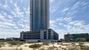 Un edificio alto y moderno con fachada curva, rodeado por un paisaje arenoso y otros edificios al fondo.
