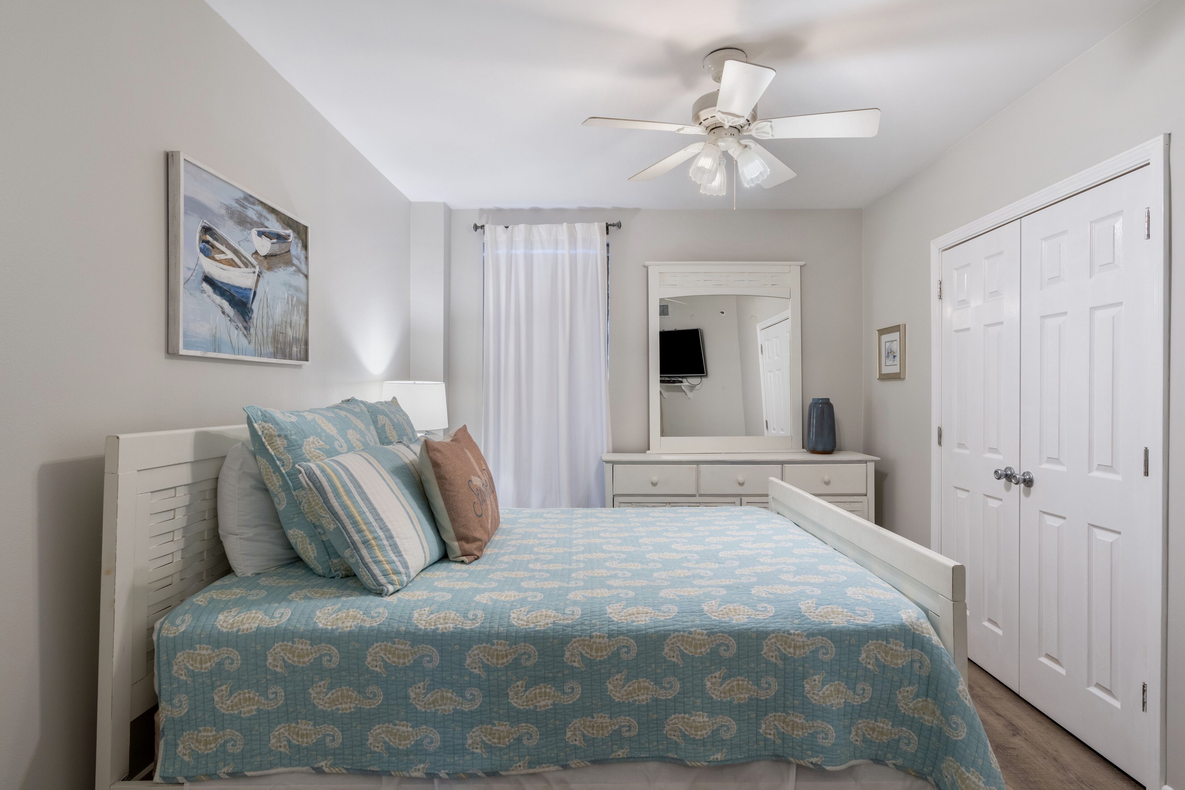 A bedroom with a bed, a ceiling fan, a painting, a dresser, a mirror, and a wardrobe.