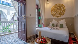 Habitación de hotel con una cama, un adorno en la pared, una mesita con flores y vistas a un balcón con plantas.
