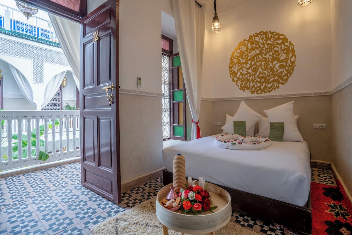 A hotel room with a bed, a decorative wall piece, a small table with flowers, and a view of a balcony with plants.
