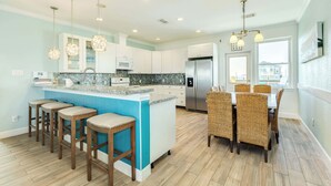 A modern kitchen with a central island, bar stools, and a dining area with wicker chairs. - Porch Paradise - 3 Beach & Water Views, Screened Porch! (Freeport)