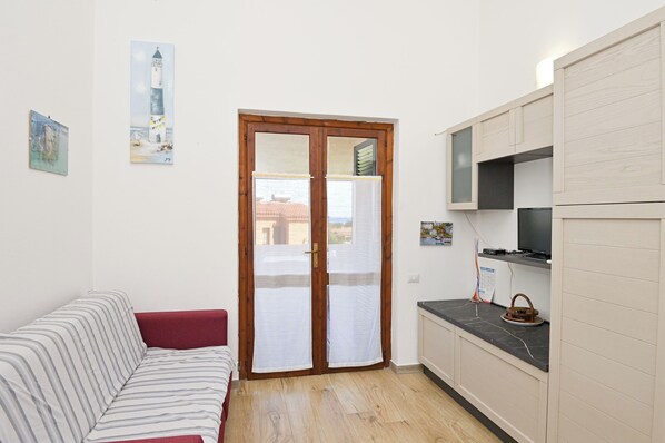 A compact living space with a sofa, a kitchen area with cabinets, a door with a view, and a painting on the wall. - AmareSardegna2 (Muravera - Costa Rei)