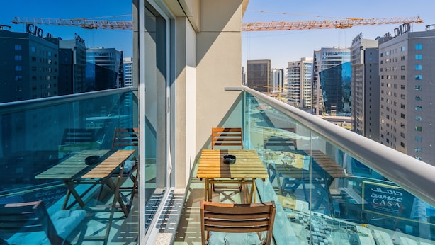 A balcony with glass railings, wooden furniture, and a city view. - Charming Studio Escape With City View (Dubai)