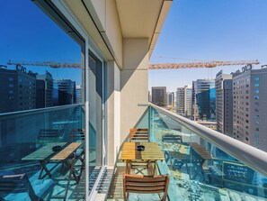 A balcony with a glass barrier, wooden furniture, and a view of city buildings.