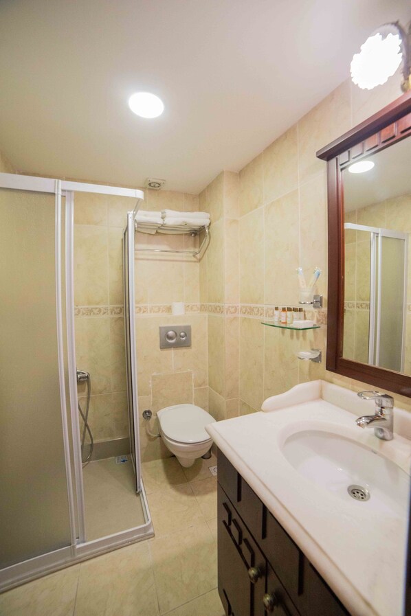 A bathroom with a glass-enclosed shower, a toilet, a sink with a mirror, and a shelf with toiletries. - Suinn Cunda (Ayvalik)