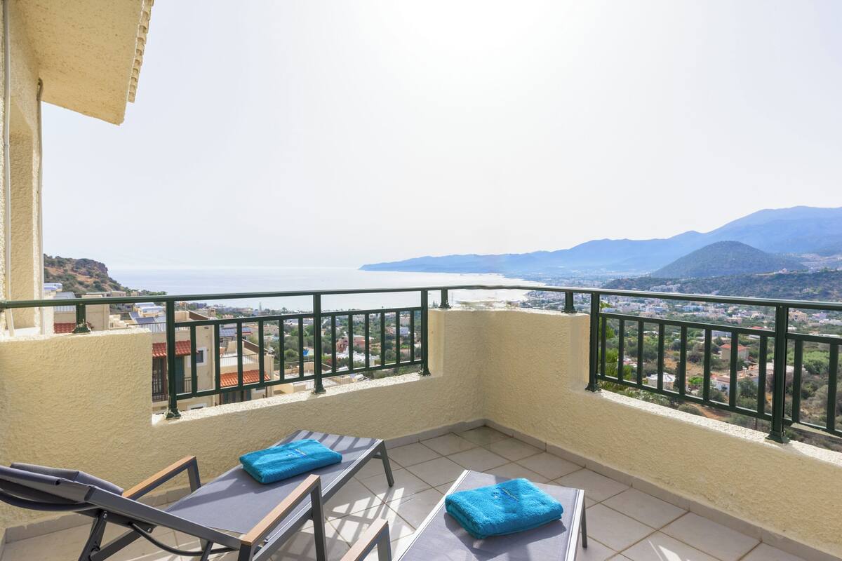 A balcony with two sun loungers and blue towels, overlooking a coastal cityscape with mountains in the distance.