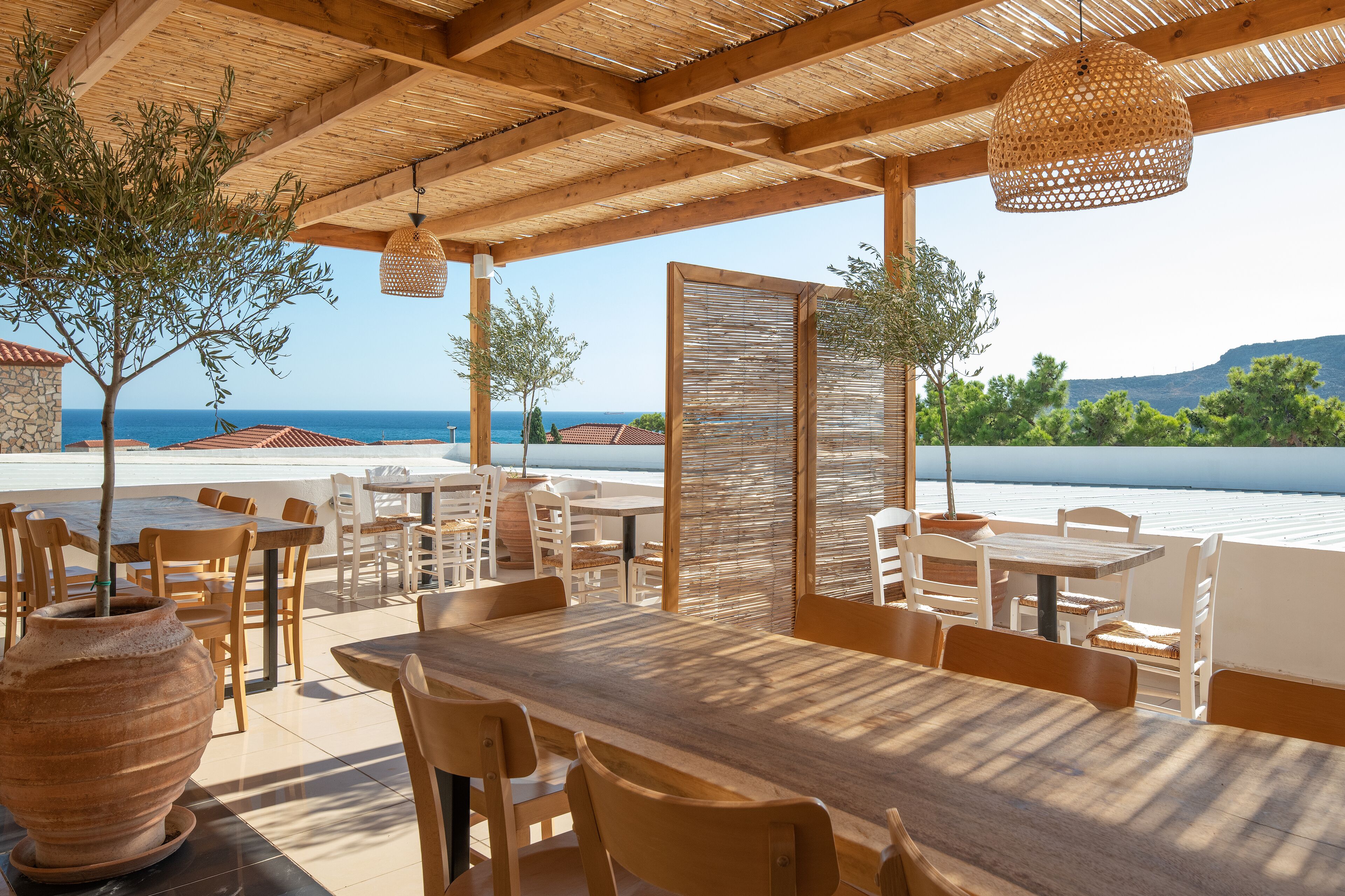 Un espace extérieur aménagé pour les repas, avec des tables et des chaises en bois, un grand pot en terre cuite et une vue sur la mer.