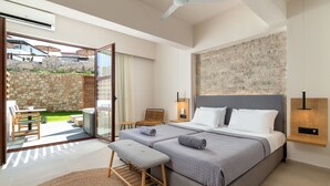 A bedroom with a large bed, a bench, a chair, and a view of a garden through an open door. - Costa Lindia Beach Resort (Rhodes)