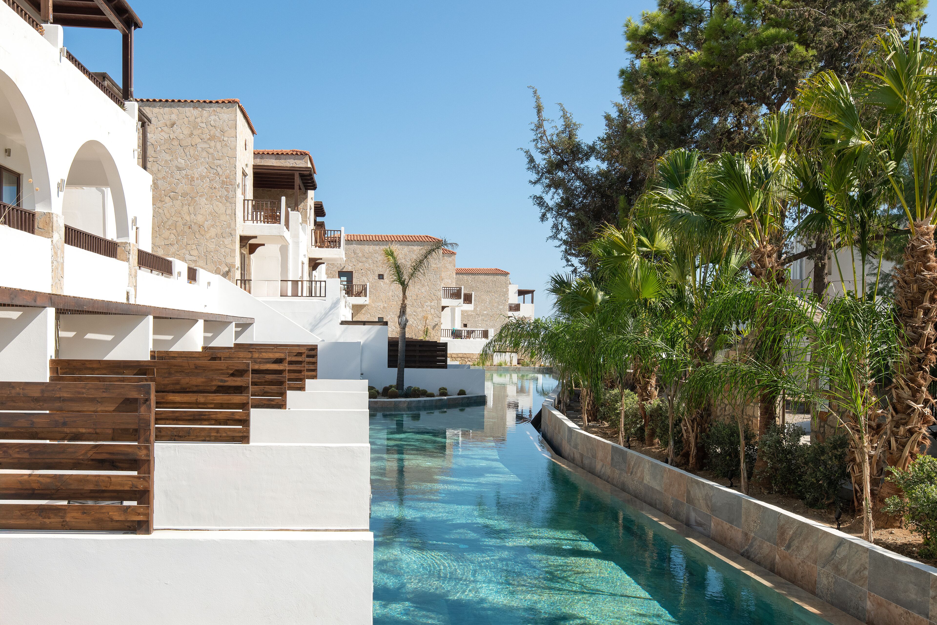 A pool area with a clear blue water feature, white buildings with stone accents, and surrounding greenery.