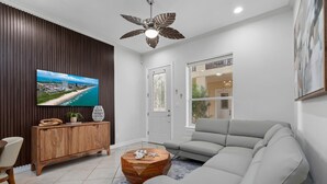 A modern living room with a sectional sofa, a wooden coffee table, a TV mounted on a wall, and a ceiling fan. - Modern Ground flr Condo 1/2 Block to Beach w Pool! (South Padre Island)