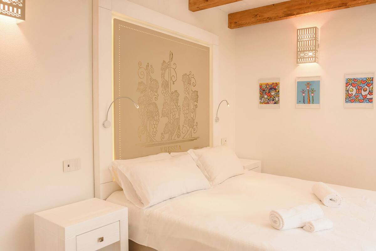 Un dormitorio con una cama, mesitas de noche blancas y una pared con un cabecero decorativo y obras de arte enmarcadas.