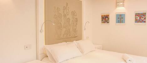 Un dormitorio con una cama, mesitas de noche blancas y una pared con un cabecero decorativo y obras de arte enmarcadas.