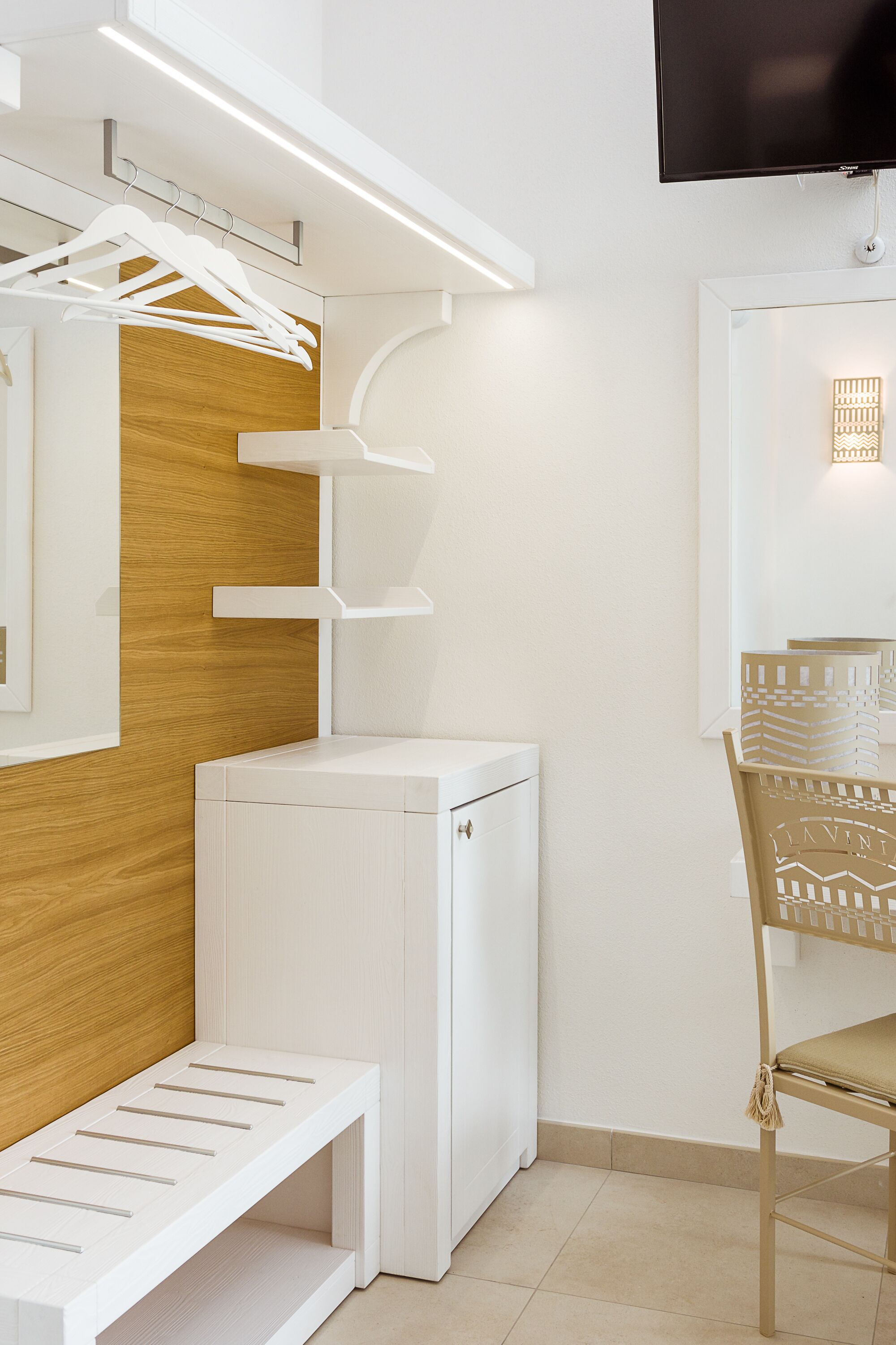 Una habitación moderna con un armario blanco, un mueble blanco pequeño, una pared de paneles de madera, un banco blanco y una silla con respaldo estampado.