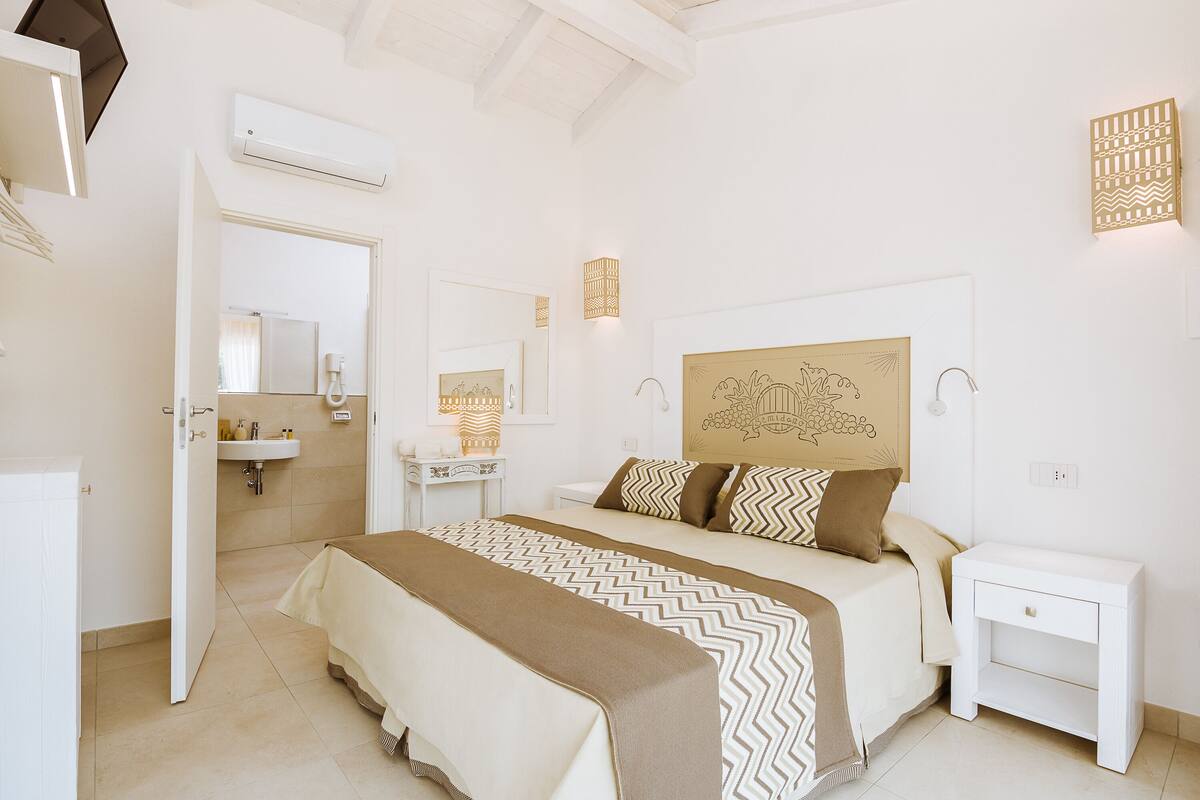 Habitación de hotel con cama, mesita de noche y baño con lavamanos y espejo.