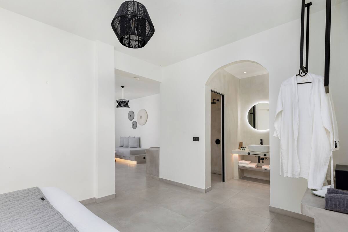 A modern hotel room with a large bed, a sitting area with a sofa and a coffee table, a bathroom with a sink and mirror, and a hallway leading to another room.