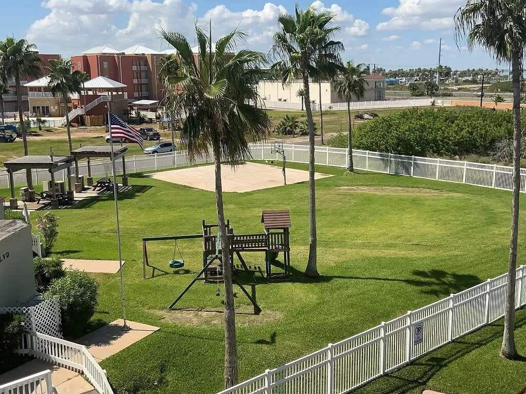 A grassy park area with playground equipment, a flag, and a white fence.