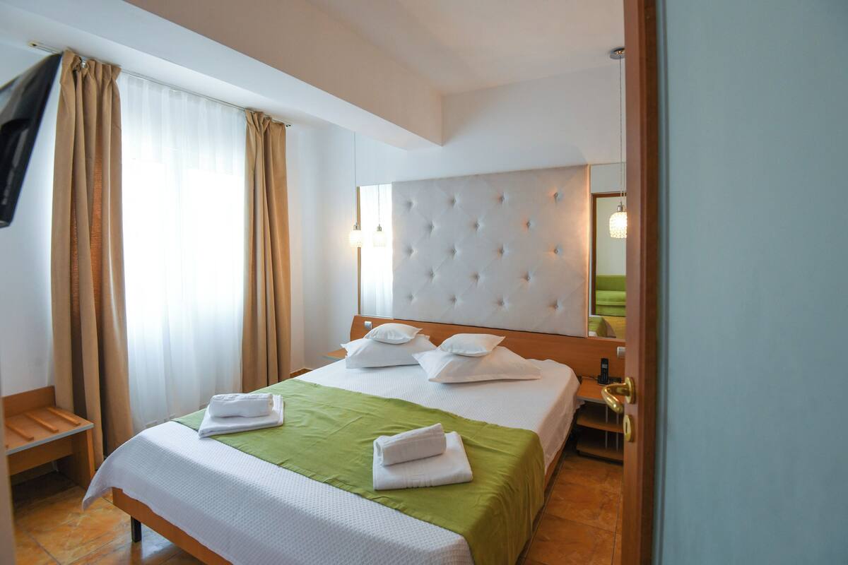 Une chambre d’hôtel avec un grand lit, une couvre-lit verte, des oreillers blancs, une tête de lit capitonnée et une table de chevet en bois avec une lampe.