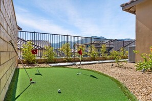 Een putting green met een vlag, omringd door een hek, bomen en gebouwen op de achtergrond.