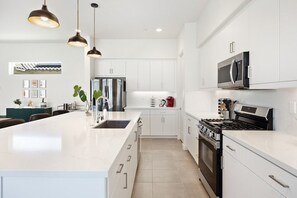 Une cuisine moderne avec des meubles blancs, un grand îlot central, des appareils électroménagers en acier inoxydable, des luminaires suspendus et un sol carrelé.