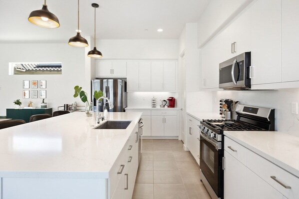 Een moderne keuken met witte kastjes, een groot eiland, roestvrijstalen apparatuur, hanglampen en een betegelde vloer.