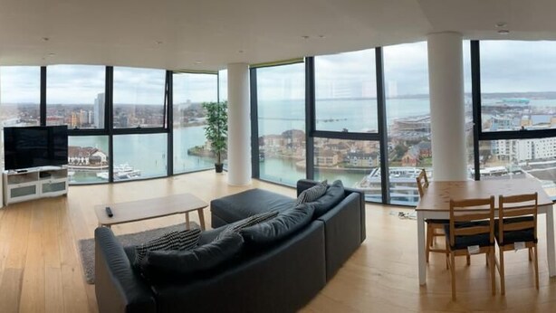 A modern living room with a large window, a black sofa, a dining table, and a kitchen area. - Stunning 3-bed, 3-bath Apartment in Southampton (Southampton)