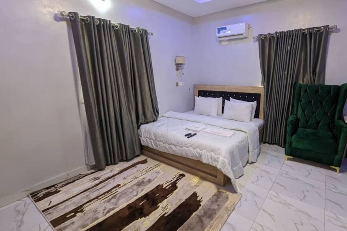A hotel room with a bed, a green armchair, a desk with a laptop, and an air conditioning unit.