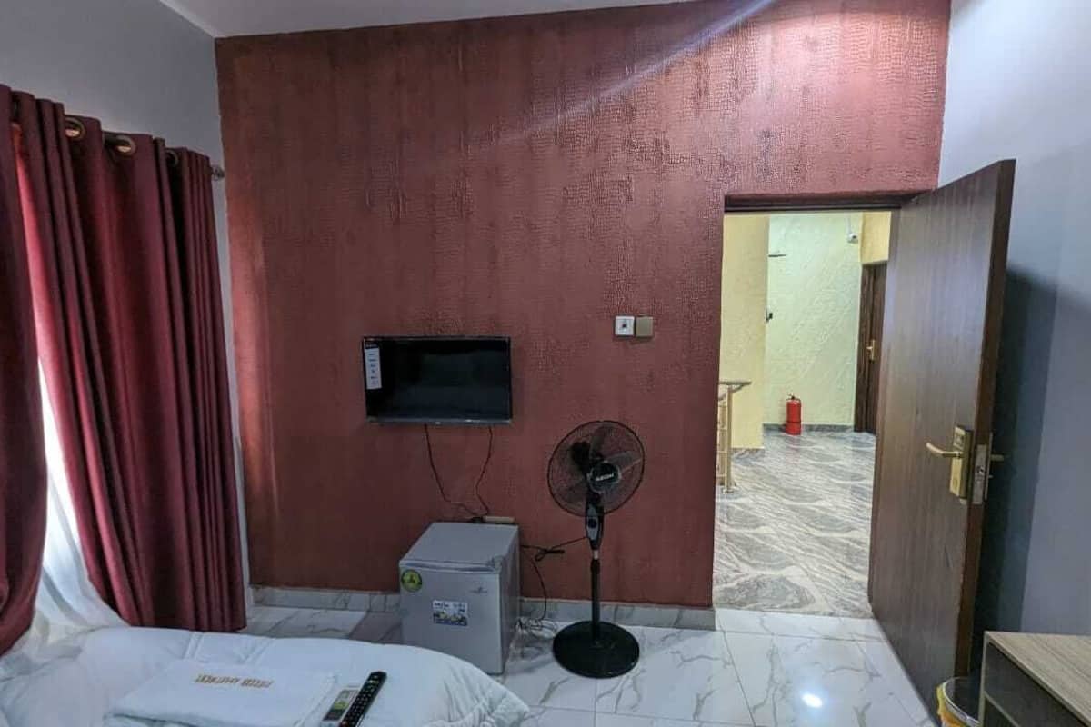A hotel room with a bed, a TV mounted on the wall, a standing fan, a small heater, and a door leading to another room.