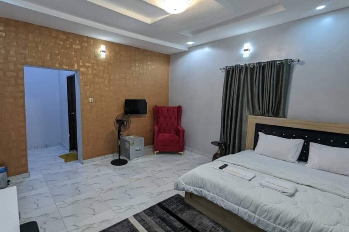 A bedroom with a bed, a red chair, a TV, and a fan.