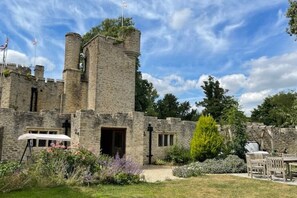 Un château en pierre doté d’une tour centrale, entouré d’un jardin où poussent différentes espèces de plantes et agrémenté d’un banc en bois.