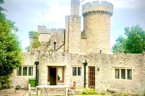 A stone castle with a round tower, a wooden door, and a small garden area with a table and chairs. - Orchardleigh Castle West, Orchardleigh Estate (Lullington)