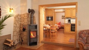 A cozy living room with a stone wall and a wood-burning stove, a dining area with a table and chairs, and a kitchen with wooden cabinets and appliances. - Vacation home, 98 sqm / sauna / 1 - 5 persons / 5 min. from the Rennsteig. (Neuhaus am Rennweg)