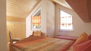 A bedroom with a wooden ceiling, a bed with a patterned bedspread, a chair, and a window with blinds. - Vacation home, 98 sqm / sauna / 1 - 5 persons / 5 min. from the Rennsteig. (Neuhaus am Rennweg)
