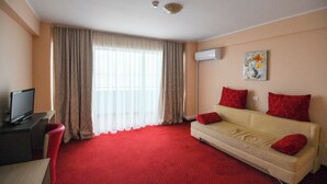 A hotel room with a red carpet, a beige sofa with red cushions, a television on a desk, and a window with sheer curtains. - Hotel Romantic (Constanta)