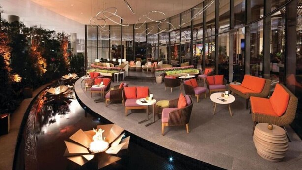 Un salón moderno con mesas redondas y sillas con cojines naranja, rodeado de paredes de vidrio y una fuente de agua.