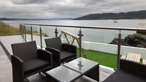A balcony with wicker furniture, a glass table, and a view of a calm lake with boats and distant hills. -  Pierside Apartments Deluxe Accommodation (Portsalon)