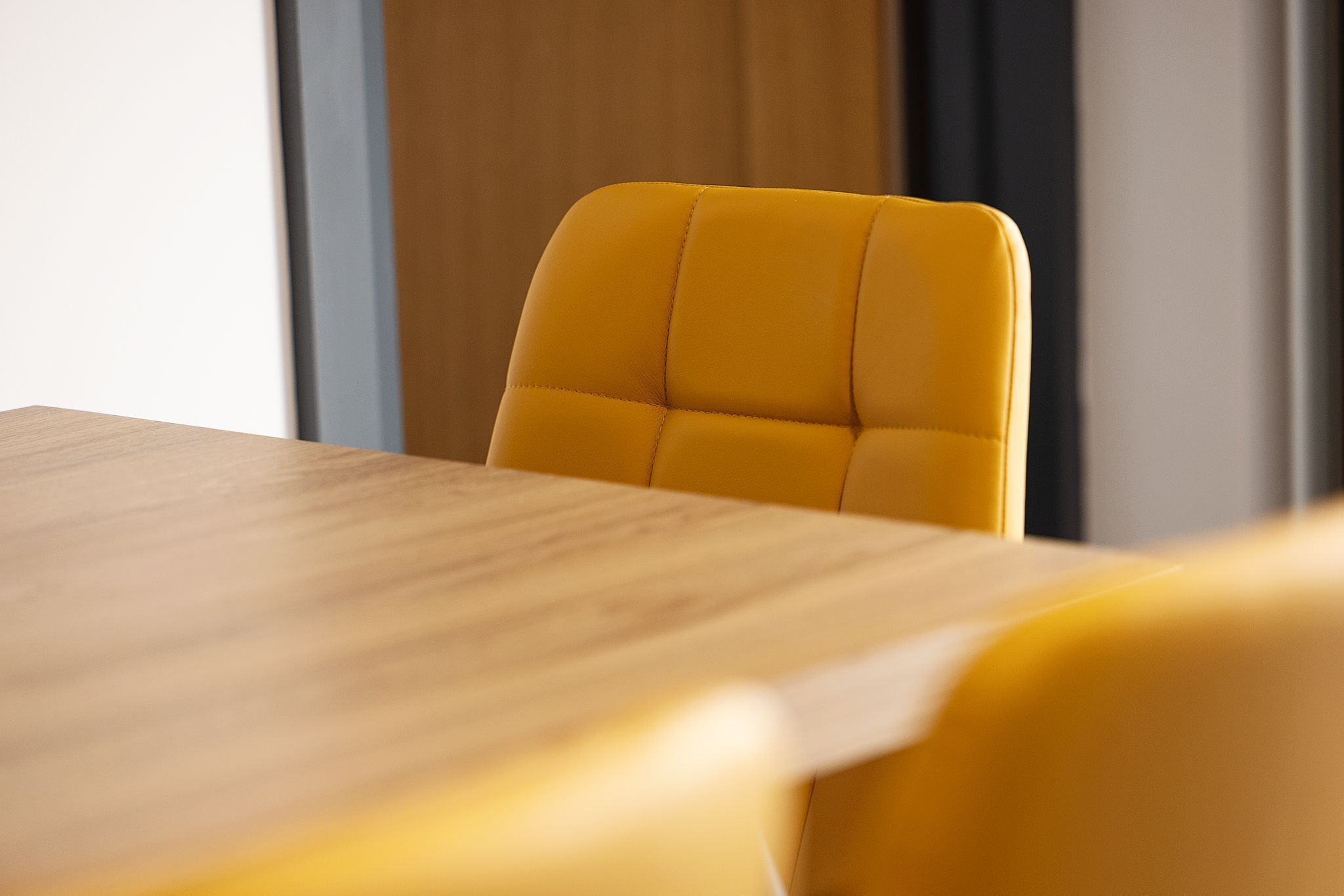 A yellow upholstered chair with a high backrest, positioned at a wooden table in a room with a neutral-toned wall and a window in the background.