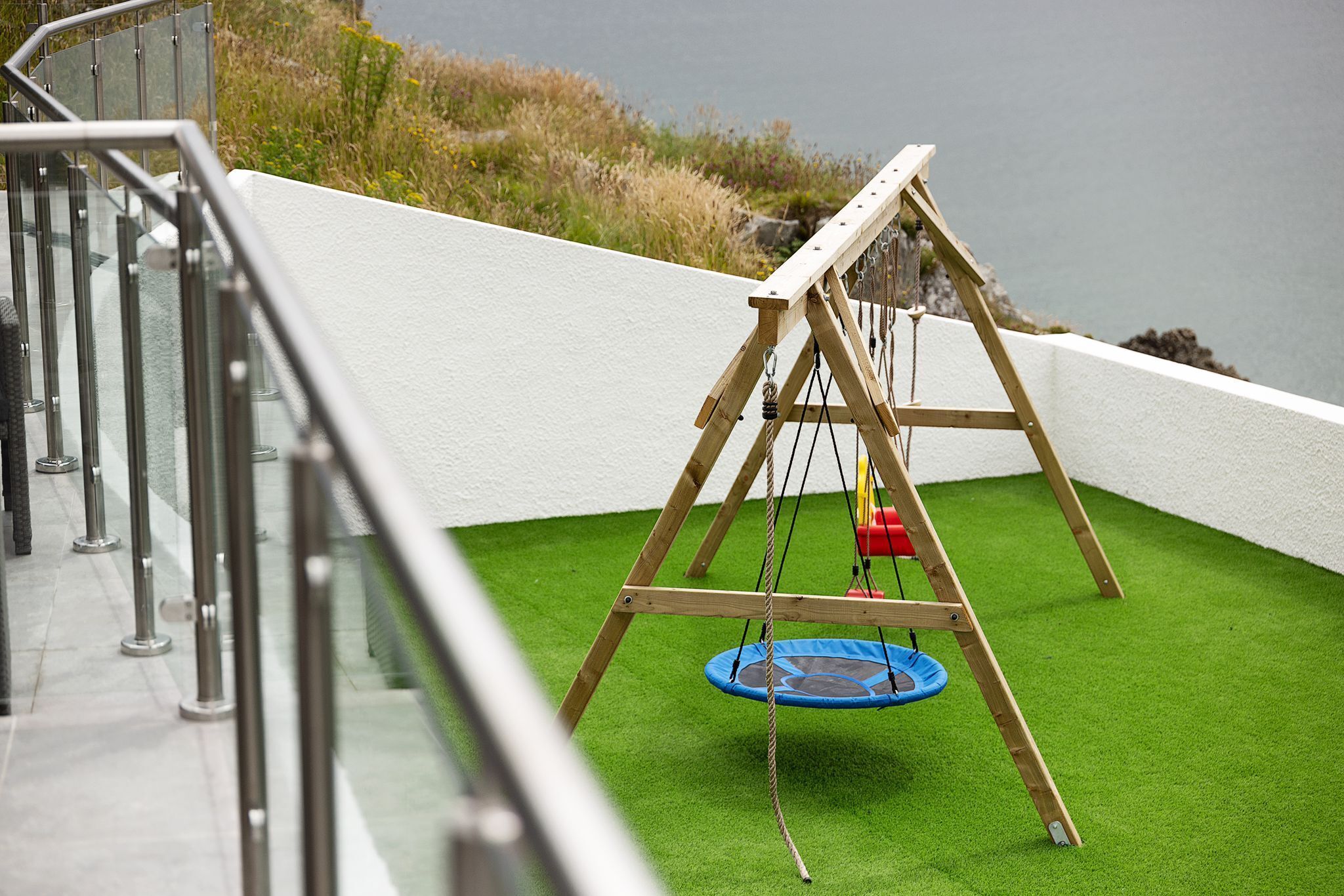 A wooden swing set with a blue trampoline on a green artificial turf, overlooking a body of water.