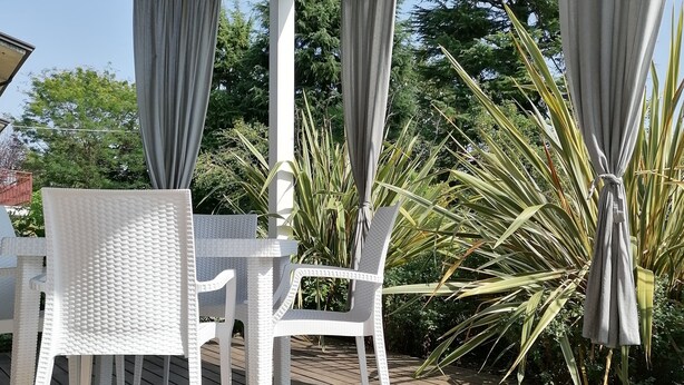 A covered outdoor area with a wooden deck, white wicker furniture, and grey curtains. - Ca' Belvedere B&B (Silea)