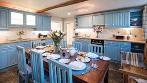 Une cuisine avec des meubles de rangement bleus, une table à manger en bois dressée pour quatre personnes et un vase rempli de fleurs posé sur la table.