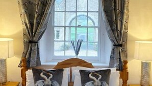Une chambre avec une grande fenêtre, des rideaux, une tête de lit en bois, deux lampes de chevet et deux coussins ornés d’un motif d’oiseau.