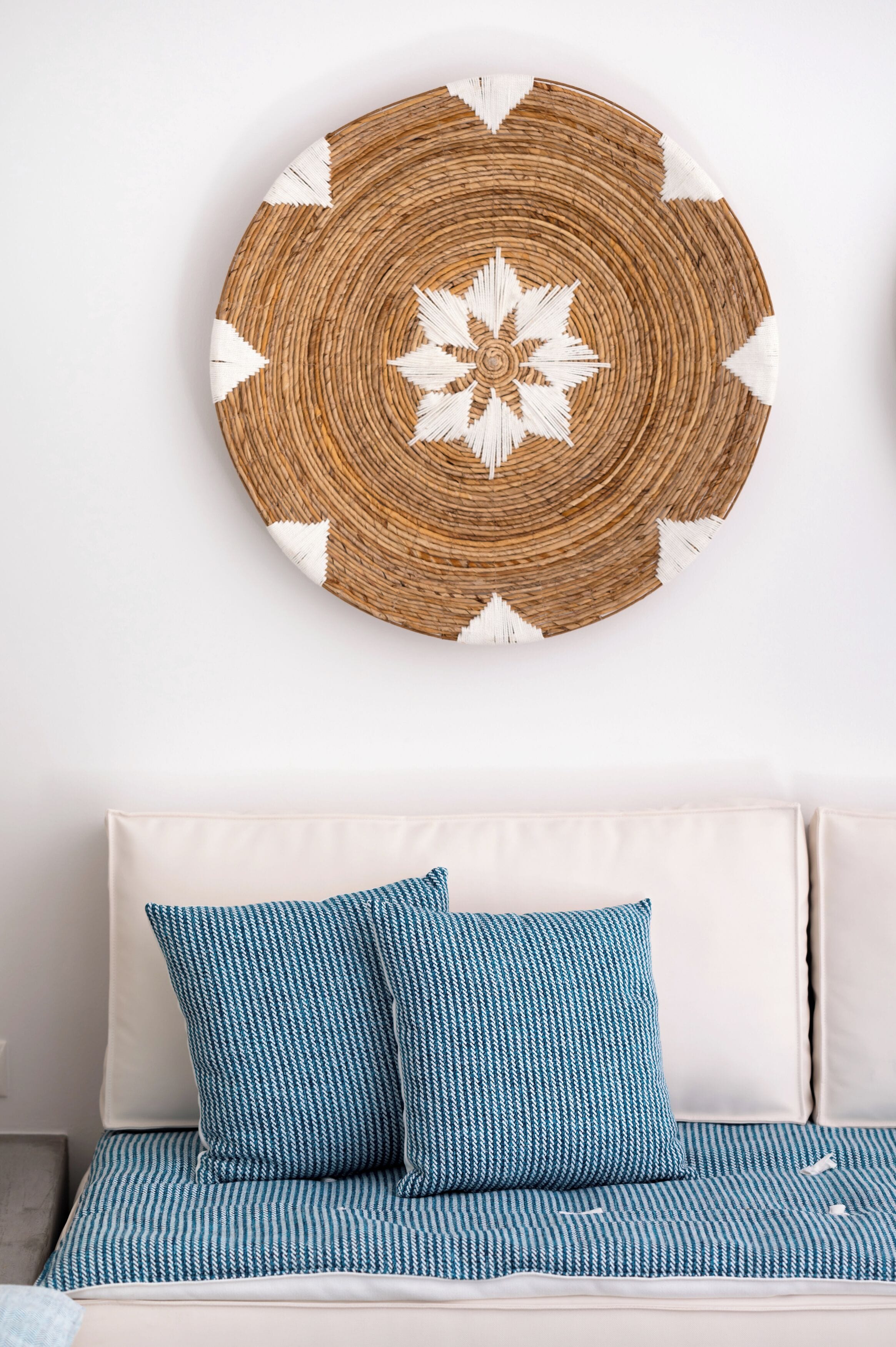 A round woven wall art with a white floral pattern, placed above a sofa with blue patterned cushions.