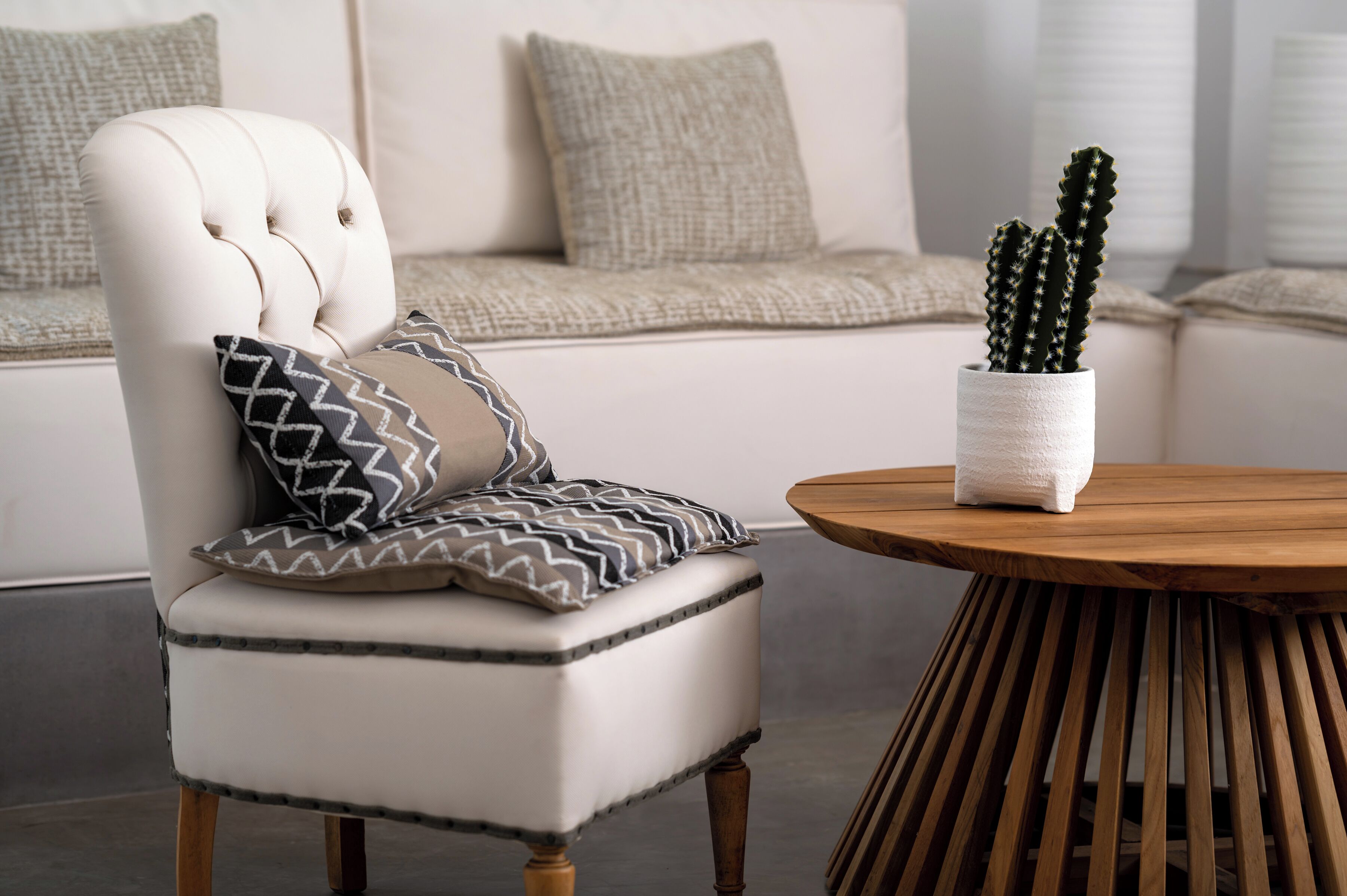A tufted armchair with patterned pillows, a round wooden table, and a cactus in a white pot.