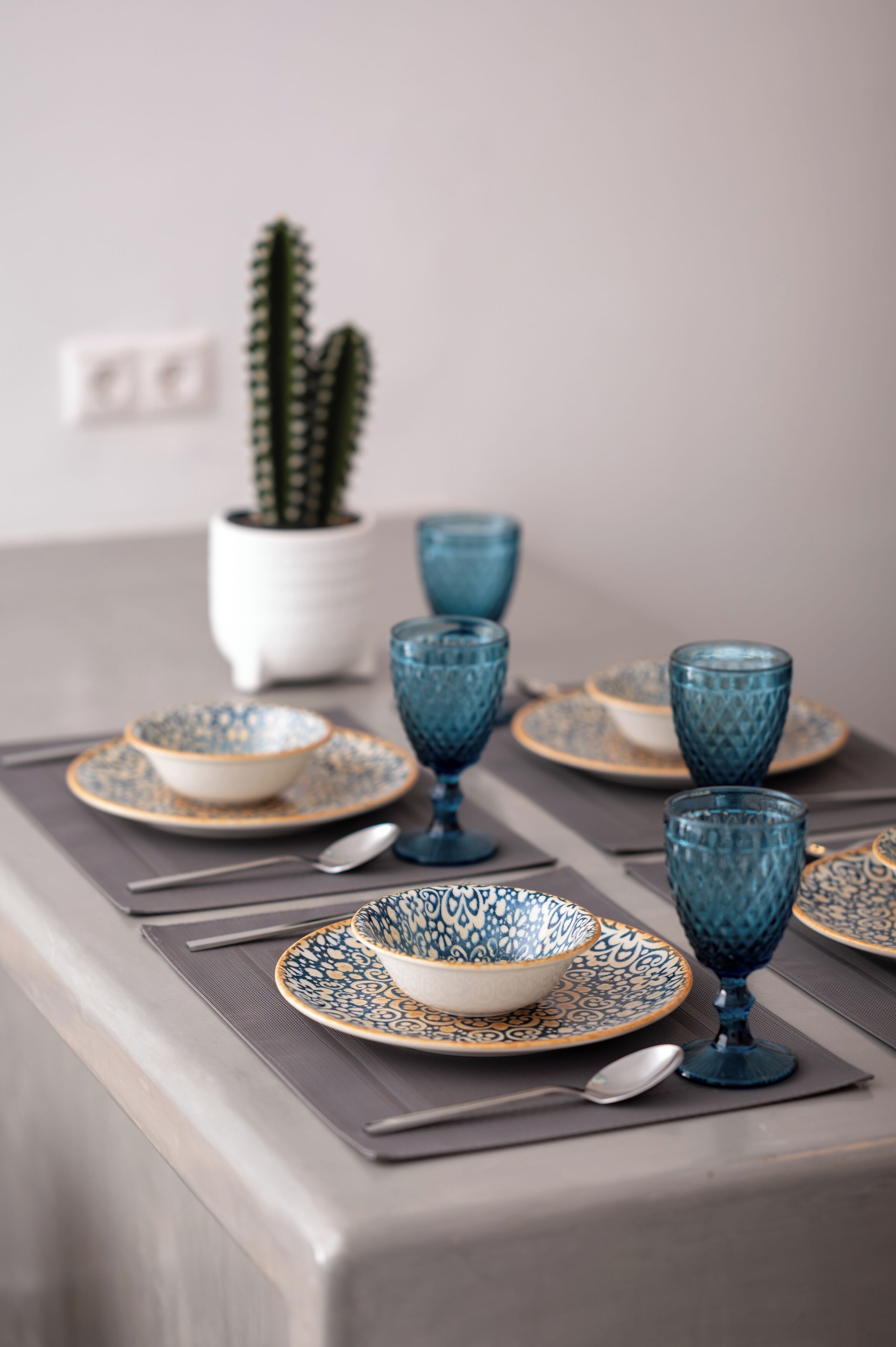 A dining table set with blue glassware and patterned plates, a cactus in a white pot, and a neutral-toned wall in the background.