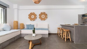A modern living room with a sofa, a round coffee table, and a kitchen area with stools. - AquaLung Suites (Santorini)