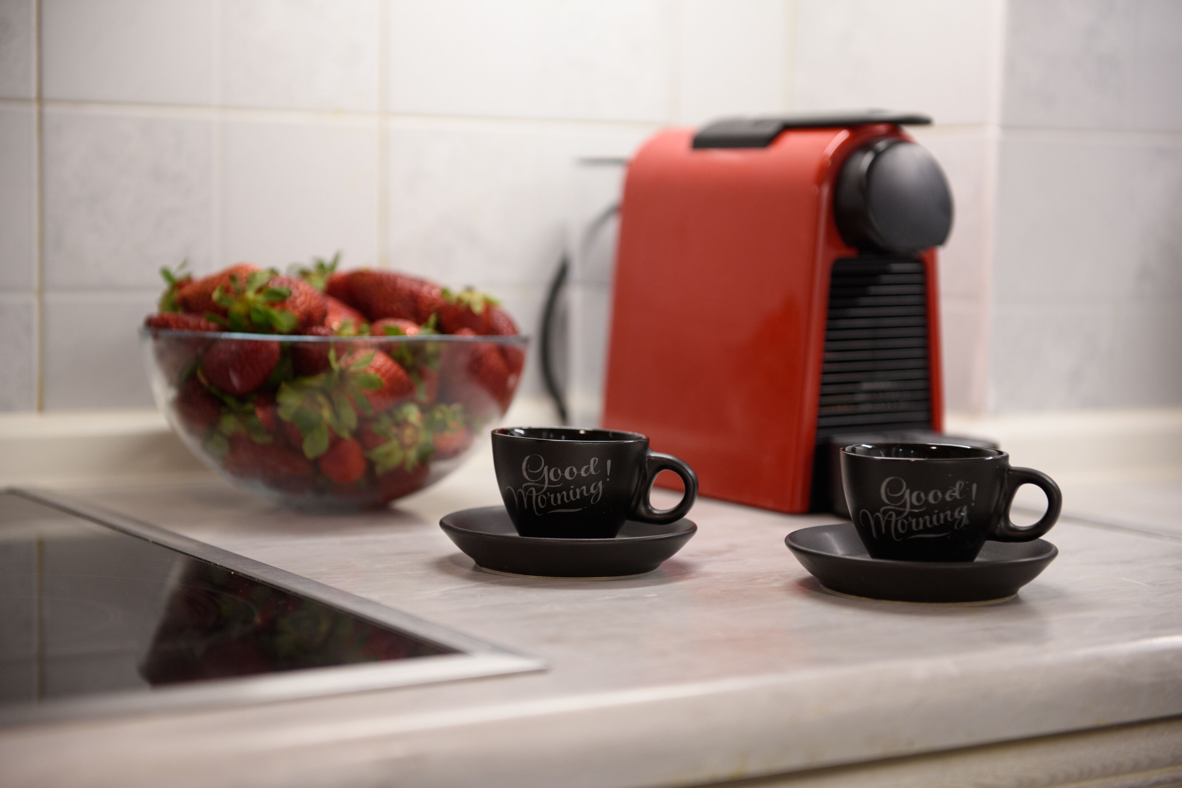 A kitchen counter with a bowl of strawberries, two black mugs with "Good Morning" text, and a red appliance.
